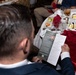 A man in a dark suit is seated at a formal banquet table, reading a printed program or brochure that appears to be for a Marine Corps Association event. The table is set with red napkins, wine glasses, dessert plates, and Asahi beer bottles, suggesting a formal military dinner or awards ceremony. A sign in the background references 'Eighth Marines Okinawa' and a 'Marine Corps Association' event.