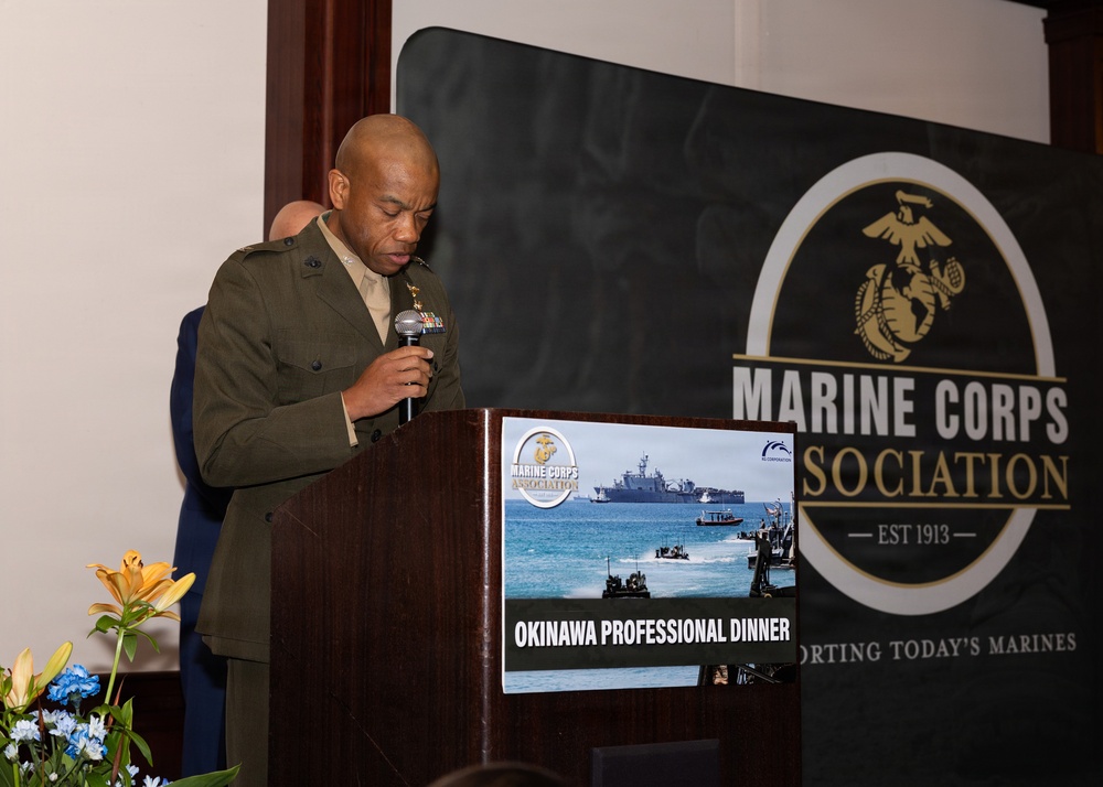A U.S. Marine Corps officer in service uniform is speaking at a podium during the Marine Corps Association Okinawa Professional Dinner. The event banner behind him displays the Marine Corps Association logo and the text 'EST 1913' along with 'Supporting Today's Marines'. A sign on the podium identifies this as the 'Okinawa Professional Dinner' and features imagery of amphibious military operations.