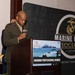A U.S. Marine Corps officer in service uniform is speaking at a podium during the Marine Corps Association Okinawa Professional Dinner. The event banner behind him displays the Marine Corps Association logo and the text 'EST 1913' along with 'Supporting Today's Marines'. A sign on the podium identifies this as the 'Okinawa Professional Dinner' and features imagery of amphibious military operations.