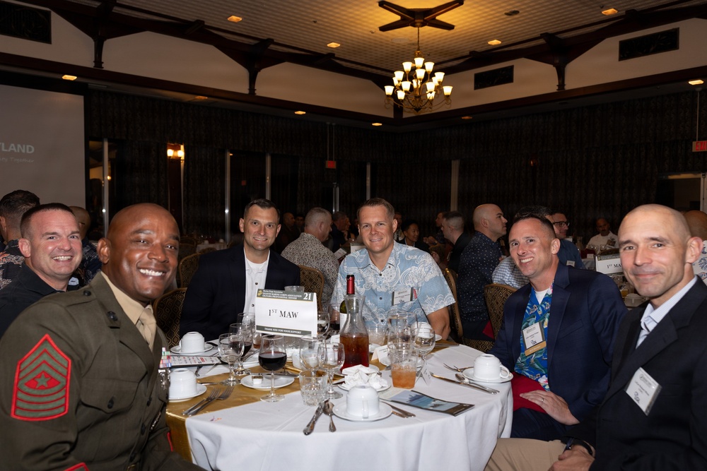 A group of approximately six men are seated at a round banquet table at what appears to be a formal military dining event. One individual is visibly wearing a U.S. Marine Corps dress uniform with sergeant major chevrons and red piping. A table placard reading '1st MAW' (1st Marine Aircraft Wing) is visible on the table.