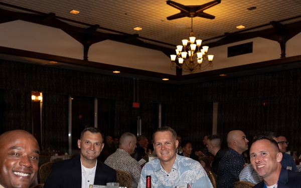 A group of approximately six men are seated at a round banquet table at what appears to be a formal military dining event. One individual is visibly wearing a U.S. Marine Corps dress uniform with sergeant major chevrons and red piping. A table placard reading '1st MAW' (1st Marine Aircraft Wing) is visible on the table.