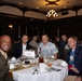 A group of approximately six men are seated at a round banquet table at what appears to be a formal military dining event. One individual is visibly wearing a U.S. Marine Corps dress uniform with sergeant major chevrons and red piping. A table placard reading '1st MAW' (1st Marine Aircraft Wing) is visible on the table.