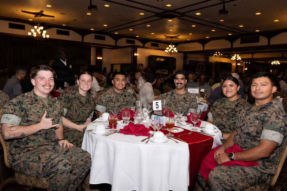Six military personnel in MARPAT camouflage uniforms are seated together at a round banquet table numbered '5', posing and smiling for the camera. The setting appears to be a formal military dining event or banquet held in a large, well-decorated hall with chandeliers and multiple occupied tables in the background. At least five appear to be U.S. Marines and one is identified as U.S. Navy based on visible uniform tape.
