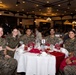 Six military personnel in MARPAT camouflage uniforms are seated together at a round banquet table numbered '5', posing and smiling for the camera. The setting appears to be a formal military dining event or banquet held in a large, well-decorated hall with chandeliers and multiple occupied tables in the background. At least five appear to be U.S. Marines and one is identified as U.S. Navy based on visible uniform tape.