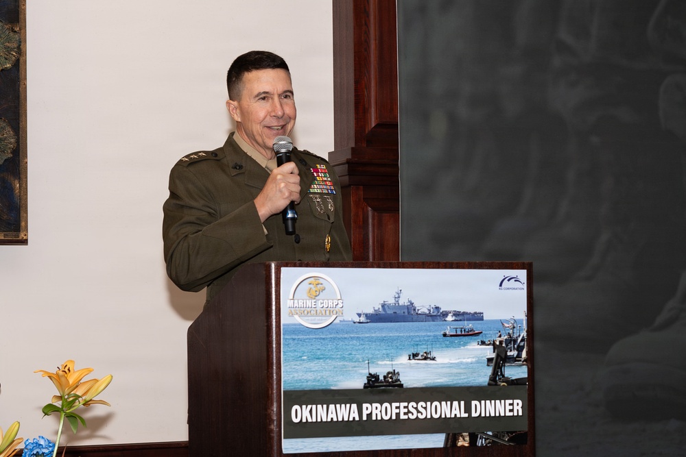 A U.S. Marine Corps officer in service uniform is speaking at a podium during the Marine Corps Association Okinawa Professional Dinner. The officer holds a microphone and stands behind a wooden lectern displaying a sign with naval amphibious operations imagery. The event appears to be a formal military professional development or networking dinner.