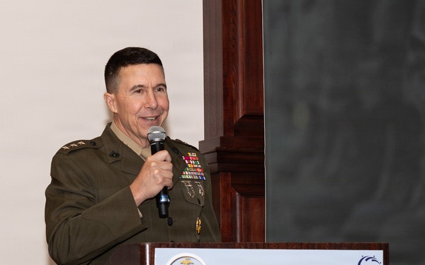 A U.S. Marine Corps officer in service uniform is speaking at a podium during the Marine Corps Association Okinawa Professional Dinner. The officer holds a microphone and stands behind a wooden lectern displaying a sign with naval amphibious operations imagery. The event appears to be a formal military professional development or networking dinner.