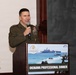 A U.S. Marine Corps officer in service uniform is speaking at a podium during the Marine Corps Association Okinawa Professional Dinner. The officer holds a microphone and stands behind a wooden lectern displaying a sign with naval amphibious operations imagery. The event appears to be a formal military professional development or networking dinner.