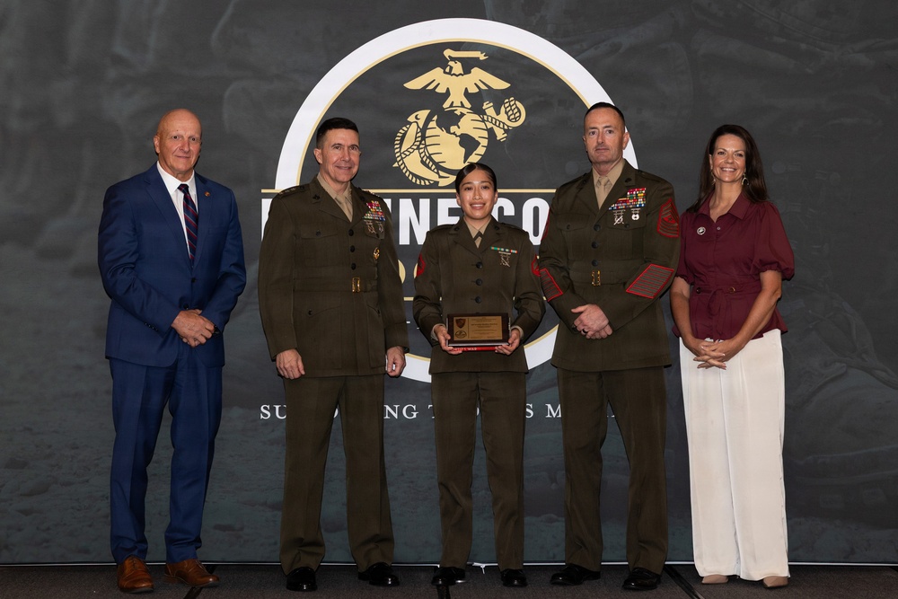 A formal U.S. Marine Corps award ceremony featuring five individuals posed in front of a backdrop displaying the Eagle, Globe, and Anchor emblem. A female Marine in the center is holding a wooden plaque/award, flanked by two senior Marines in uniform and two civilians in business attire. The setting appears to be an official recognition event.