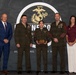 A formal U.S. Marine Corps award ceremony featuring five individuals posed in front of a backdrop displaying the Eagle, Globe, and Anchor emblem. A female Marine in the center is holding a wooden plaque/award, flanked by two senior Marines in uniform and two civilians in business attire. The setting appears to be an official recognition event.