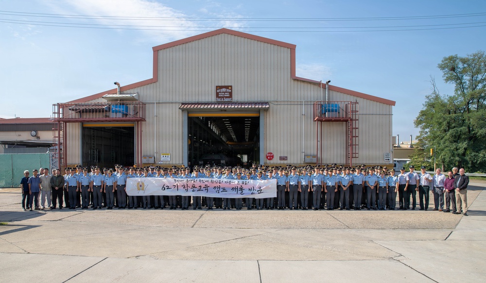 A large group of uniformed personnel, appearing to be military or law enforcement trainees, pose for a formal group photograph in front of an industrial warehouse or maintenance facility. They are holding a banner with Korean text that reads '62기 학동교육 캠프 개울 받으' (roughly translating to a 62nd class training camp completion/welcome). A smaller group of civilians in plain clothes stands to the right side of the formation.