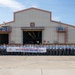 A large group of uniformed personnel, appearing to be military or law enforcement trainees, pose for a formal group photograph in front of an industrial warehouse or maintenance facility. They are holding a banner with Korean text that reads '62기 학동교육 캠프 개울 받으' (roughly translating to a 62nd class training camp completion/welcome). A smaller group of civilians in plain clothes stands to the right side of the formation.