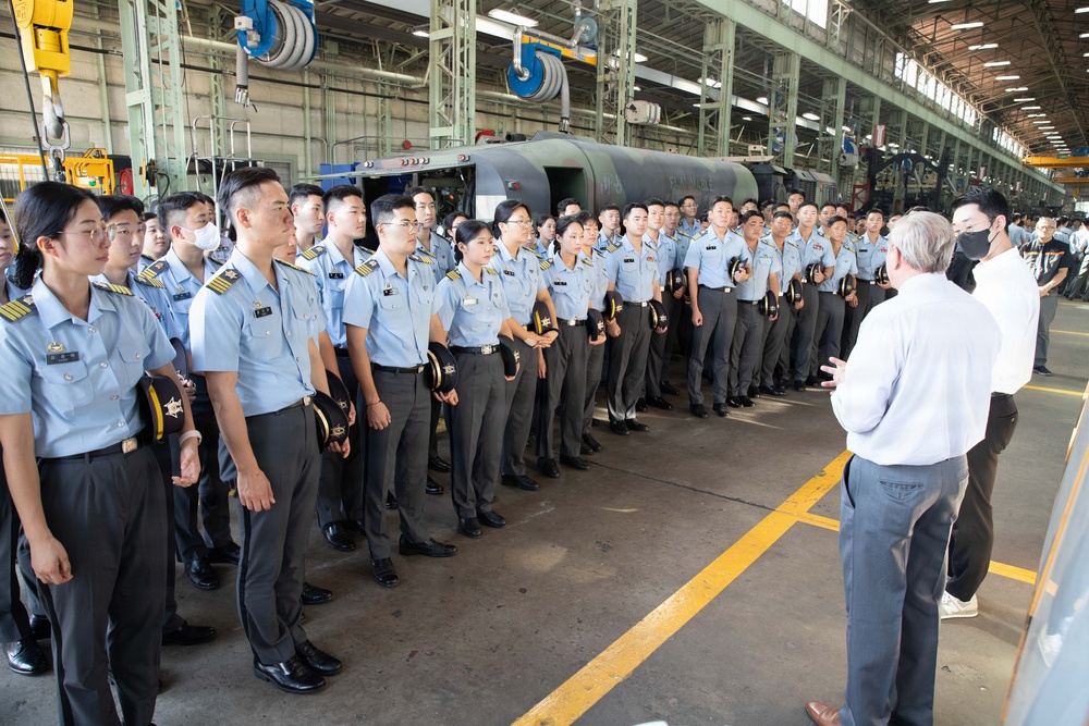 A large group of uniformed military cadets or officers, appearing to be from an East Asian nation, are assembled in a row inside an industrial facility or hangar. Two civilians in casual attire are addressing or briefing the group. The cadets are wearing light blue uniforms with dark trousers and are holding their caps.