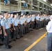 A large group of uniformed military cadets or officers, appearing to be from an East Asian nation, are assembled in a row inside an industrial facility or hangar. Two civilians in casual attire are addressing or briefing the group. The cadets are wearing light blue uniforms with dark trousers and are holding their caps.