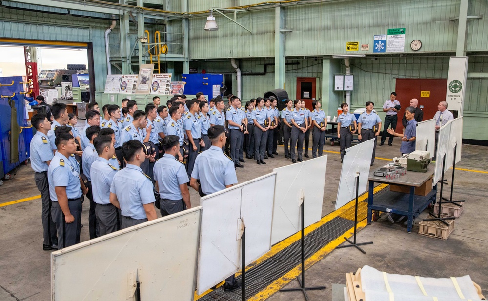 A large group of uniformed military or cadet personnel, appearing to be of Asian origin, are assembled in an industrial facility or maintenance hangar. They are being addressed by two or three civilian or senior personnel standing near whiteboards and a table with equipment. The setting appears to be a formal briefing or orientation session.