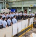A large group of uniformed military or cadet personnel, appearing to be of Asian origin, are assembled in an industrial facility or maintenance hangar. They are being addressed by two or three civilian or senior personnel standing near whiteboards and a table with equipment. The setting appears to be a formal briefing or orientation session.
