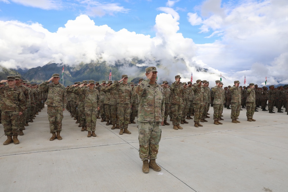A large military formation of soldiers in camouflage uniforms is performing a salute during what appears to be a formal ceremony or parade. The soldiers are arranged in organized rows on a concrete parade ground, with mountainous terrain and dramatic cloud formations visible in the background. Multiple national flags are visible among the formation, suggesting this is a multinational military exercise or joint ceremony.