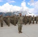 A large military formation of soldiers in camouflage uniforms is performing a salute during what appears to be a formal ceremony or parade. The soldiers are arranged in organized rows on a concrete parade ground, with mountainous terrain and dramatic cloud formations visible in the background. Multiple national flags are visible among the formation, suggesting this is a multinational military exercise or joint ceremony.