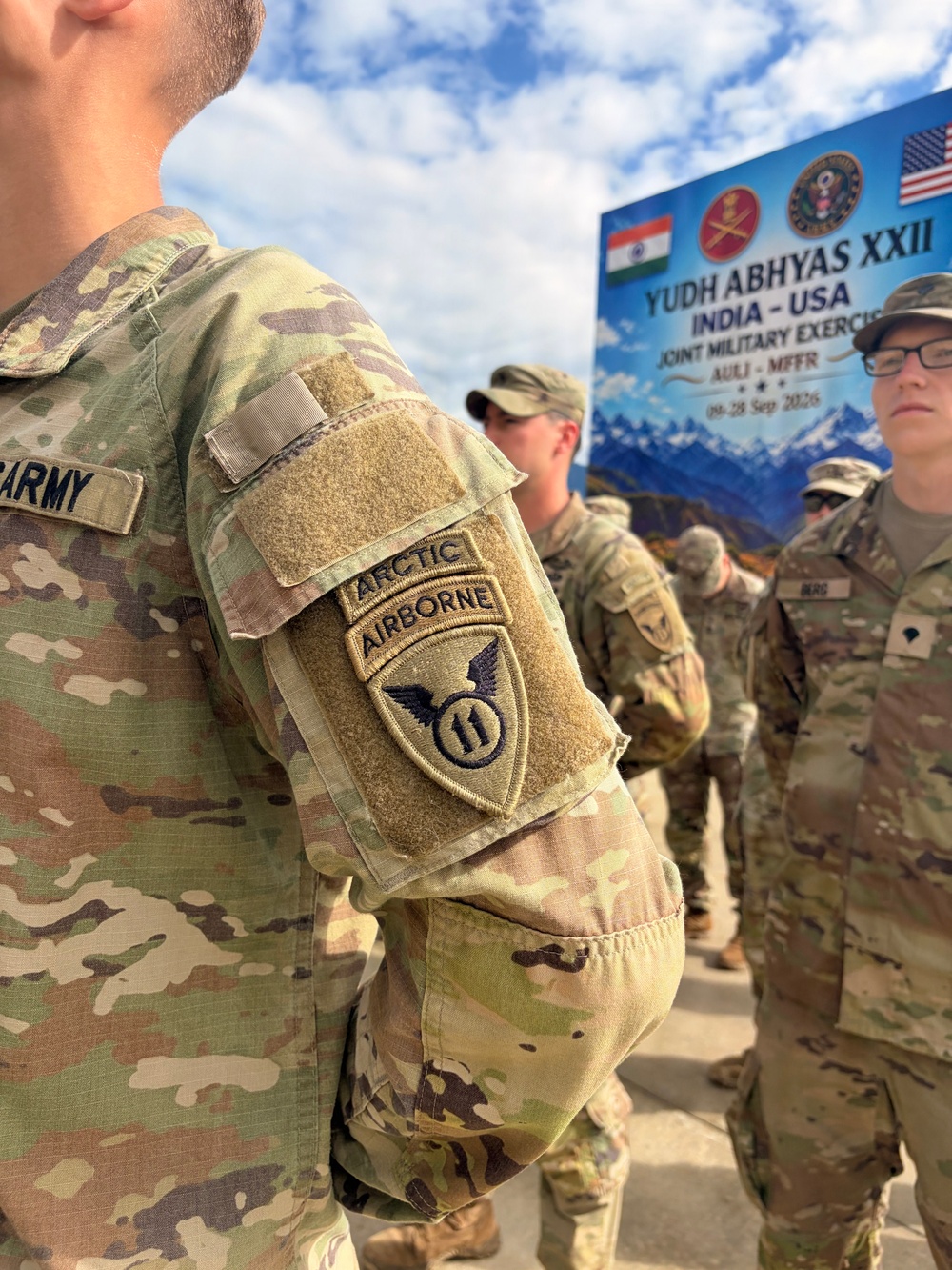 US Army soldiers in MultiCam/OCP camouflage uniforms are gathered at the Yudh Abhyas XXII India-USA Joint Military Exercise. The foreground soldier displays an '11th Airborne Division' patch with 'Arctic' and 'Airborne' tabs on the left sleeve. A banner in the background identifies the exercise as taking place 19-28 September 2023 at AULI-MFFR.