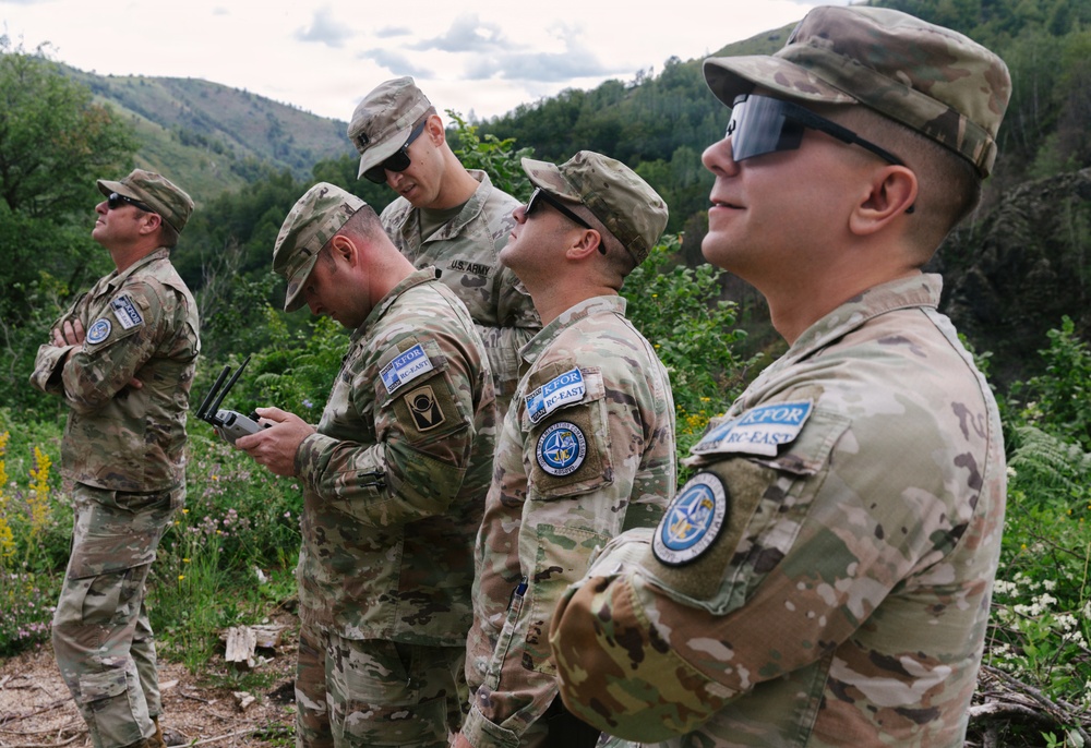 A group of five U.S. Army soldiers in MultiCam camouflage uniforms are standing outdoors in a wooded, hilly terrain. They are wearing patches identifying them as part of KFOR (Kosovo Force) RC-East, a NATO-led peacekeeping mission. The soldiers appear to be observing or surveying their surroundings during what appears to be a patrol or reconnaissance activity.