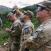 A group of five U.S. Army soldiers in MultiCam camouflage uniforms are standing outdoors in a wooded, hilly terrain. They are wearing patches identifying them as part of KFOR (Kosovo Force) RC-East, a NATO-led peacekeeping mission. The soldiers appear to be observing or surveying their surroundings during what appears to be a patrol or reconnaissance activity.