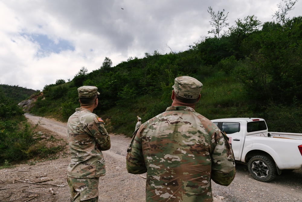 Two U.S. Army soldiers in MultiCam/OCP camouflage uniforms are standing on a gravel road, facing away from the camera and looking toward the sky or down a wooded hillside path. One soldier has the name tape 'HUCK' visible on their uniform, and both wear American flag patches on their sleeves. A white pickup truck is parked to their right, and the setting appears to be a rural, forested area.