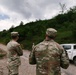 Two U.S. Army soldiers in MultiCam/OCP camouflage uniforms are standing on a gravel road, facing away from the camera and looking toward the sky or down a wooded hillside path. One soldier has the name tape 'HUCK' visible on their uniform, and both wear American flag patches on their sleeves. A white pickup truck is parked to their right, and the setting appears to be a rural, forested area.