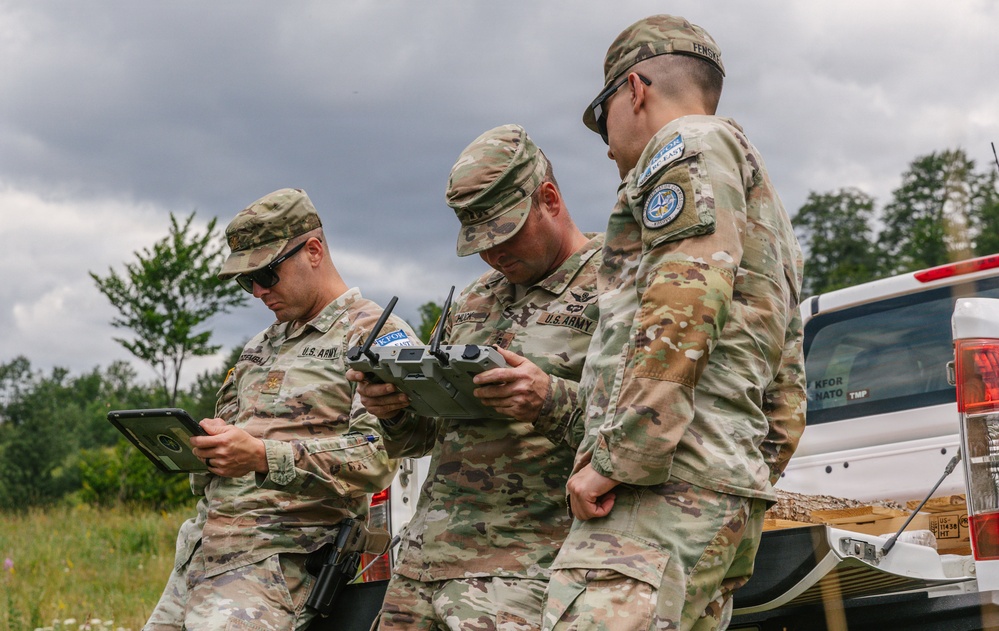 Three U.S. Army soldiers in MultiCam/OCP camouflage uniforms are operating what appears to be a drone controller and tablet device in an outdoor field setting. They are associated with NATO's KFOR (Kosovo Force) mission, as indicated by patches and vehicle markings visible in the image. The soldiers appear to be conducting UAS (Unmanned Aerial System) operations or training.