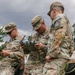 Three U.S. Army soldiers in MultiCam/OCP camouflage uniforms are operating what appears to be a drone controller and tablet device in an outdoor field setting. They are associated with NATO's KFOR (Kosovo Force) mission, as indicated by patches and vehicle markings visible in the image. The soldiers appear to be conducting UAS (Unmanned Aerial System) operations or training.