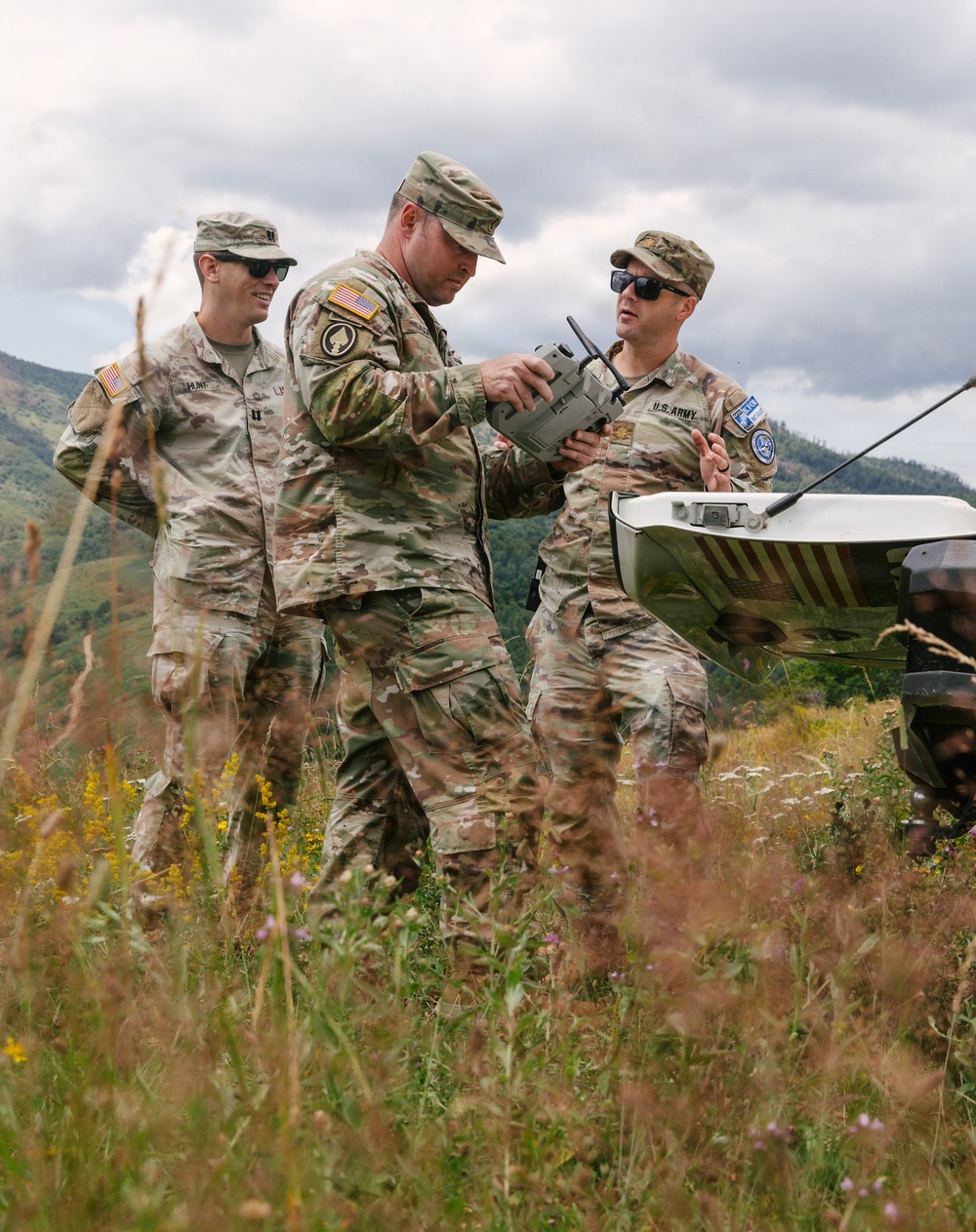 Three U.S. Army soldiers in MultiCam/OCP camouflage uniforms are gathered in an outdoor field environment, examining what appears to be a drone remote control or ground control station. They are standing among tall grass and wildflowers with mountainous terrain visible in the background. The central soldier is holding the controller while the soldier on the right appears to be providing instruction or explanation.