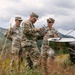 Three U.S. Army soldiers in MultiCam/OCP camouflage uniforms are gathered in an outdoor field environment, examining what appears to be a drone remote control or ground control station. They are standing among tall grass and wildflowers with mountainous terrain visible in the background. The central soldier is holding the controller while the soldier on the right appears to be providing instruction or explanation.