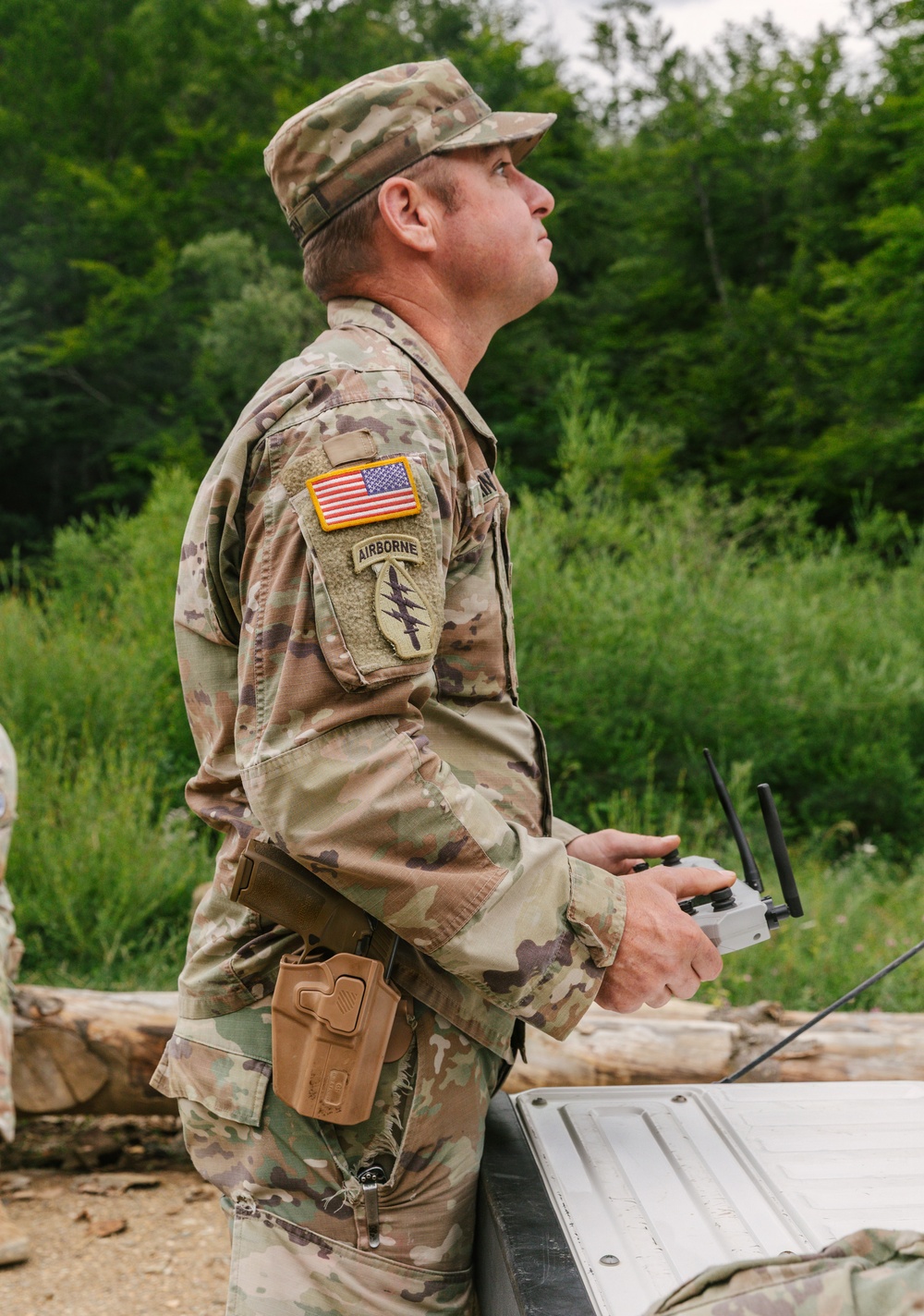 A U.S. Army soldier in Operational Camouflage Pattern (OCP) uniform is operating a drone remote controller while looking upward, likely tracking an unmanned aerial vehicle in flight. The soldier wears an Airborne tab and a unit patch with lightning bolts on the right sleeve, along with a U.S. flag patch. The setting appears to be an outdoor training or operational environment surrounded by dense vegetation.