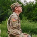 A U.S. Army soldier in Operational Camouflage Pattern (OCP) uniform is operating a drone remote controller while looking upward, likely tracking an unmanned aerial vehicle in flight. The soldier wears an Airborne tab and a unit patch with lightning bolts on the right sleeve, along with a U.S. flag patch. The setting appears to be an outdoor training or operational environment surrounded by dense vegetation.