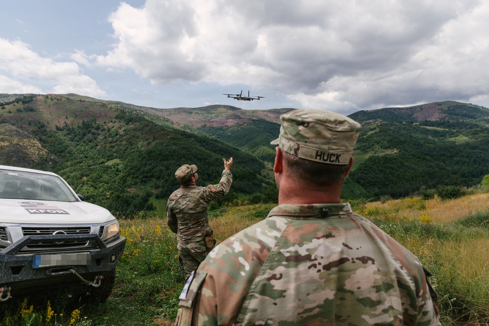 Two soldiers in camouflage uniforms observe or direct a twin-engine military aircraft flying low over a mountainous, forested landscape. A white vehicle marked 'KFOR' (Kosovo Force, NATO peacekeeping mission) is visible to the left. One soldier, identified by name tape as 'HUCK', watches from the foreground while another gestures toward the aircraft.