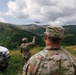Two soldiers in camouflage uniforms observe or direct a twin-engine military aircraft flying low over a mountainous, forested landscape. A white vehicle marked 'KFOR' (Kosovo Force, NATO peacekeeping mission) is visible to the left. One soldier, identified by name tape as 'HUCK', watches from the foreground while another gestures toward the aircraft.