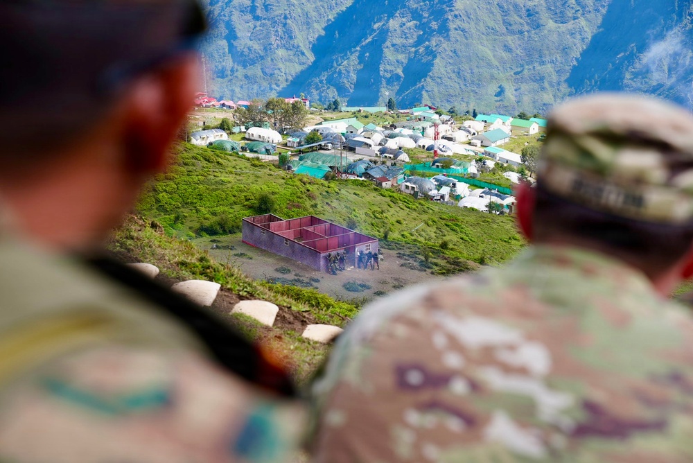 Two military personnel in camouflage uniforms are observing a military training camp or base situated on a hillside in a mountainous region. The camp contains numerous white and teal-roofed structures, tents, and what appears to be a mock building used for tactical training exercises. The foreground figures are out of focus, drawing attention to the camp and mountain backdrop.