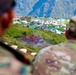Two military personnel in camouflage uniforms are observing a military training camp or base situated on a hillside in a mountainous region. The camp contains numerous white and teal-roofed structures, tents, and what appears to be a mock building used for tactical training exercises. The foreground figures are out of focus, drawing attention to the camp and mountain backdrop.