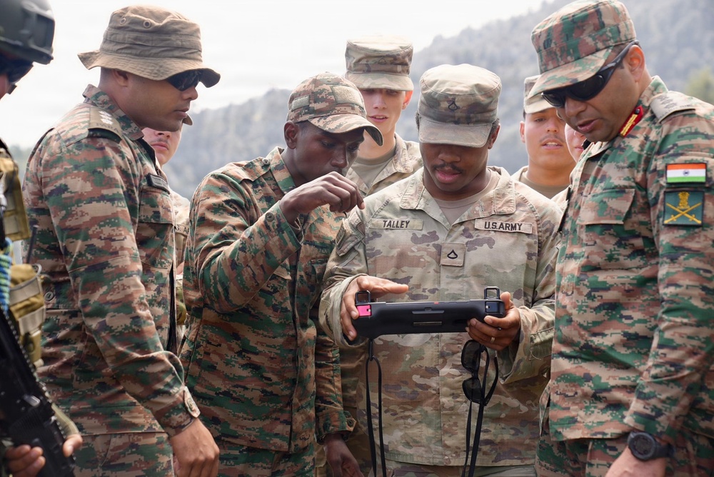 The image shows a joint military exercise between U.S. Army soldiers and Indian Army personnel. A U.S. Army soldier named Talley is holding a tactical tablet/device while personnel from both forces gather around to examine it. The interaction appears to be a demonstration or briefing involving digital equipment, likely during a bilateral training exercise.