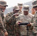The image shows a joint military exercise between U.S. Army soldiers and Indian Army personnel. A U.S. Army soldier named Talley is holding a tactical tablet/device while personnel from both forces gather around to examine it. The interaction appears to be a demonstration or briefing involving digital equipment, likely during a bilateral training exercise.
