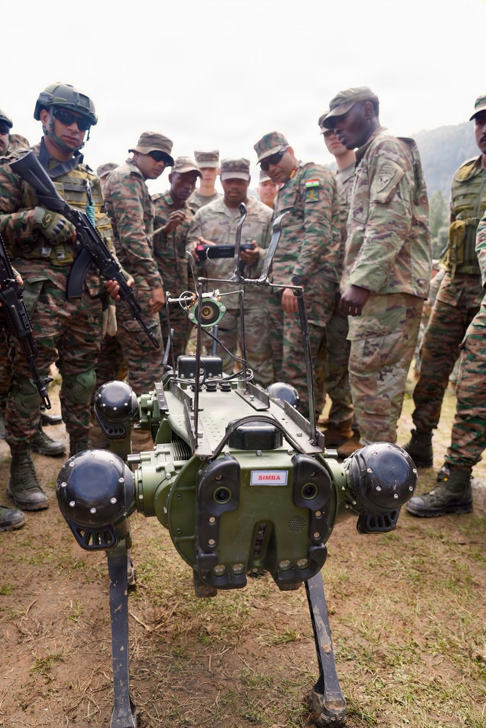 A group of military personnel in mixed camouflage uniforms are gathered around a quadruped military robot labeled 'SIMBA'. The soldiers appear to represent at least two different nations, identifiable by distinct camouflage patterns including Indian Army DPM and US Army OCP uniforms. The scene appears to be a demonstration or training exercise conducted in an outdoor, hilly terrain.