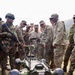 A group of military personnel in mixed camouflage uniforms are gathered around a quadruped military robot labeled 'SIMBA'. The soldiers appear to represent at least two different nations, identifiable by distinct camouflage patterns including Indian Army DPM and US Army OCP uniforms. The scene appears to be a demonstration or training exercise conducted in an outdoor, hilly terrain.