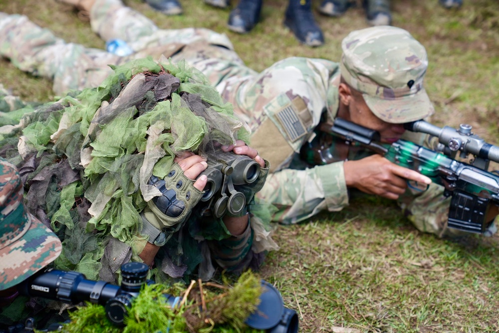 Military personnel are engaged in a sniper or reconnaissance training exercise in an outdoor field setting. One soldier wearing a ghillie suit is using binoculars for observation, while another soldier in MultiCam/OCP uniform with a US flag patch is aiming a scoped rifle. Additional personnel and equipment are visible in the background and foreground.
