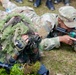 Military personnel are engaged in a sniper or reconnaissance training exercise in an outdoor field setting. One soldier wearing a ghillie suit is using binoculars for observation, while another soldier in MultiCam/OCP uniform with a US flag patch is aiming a scoped rifle. Additional personnel and equipment are visible in the background and foreground.