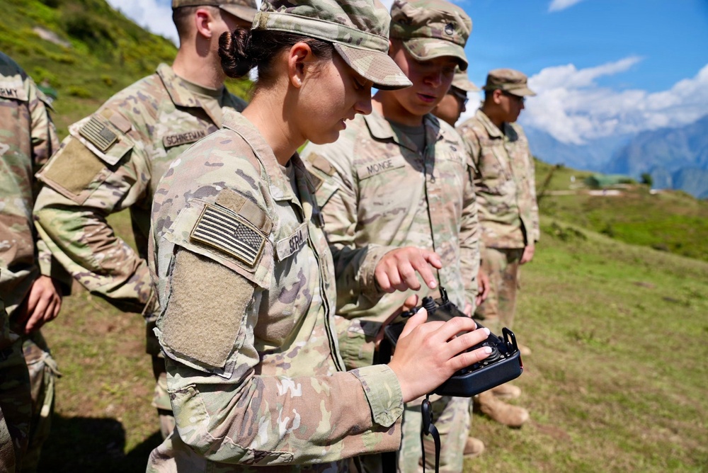 A group of U.S. Army soldiers in MultiCam (OCP) camouflage uniforms are gathered outdoors in a mountainous, green terrain under a partly cloudy sky. A female soldier in the foreground is operating what appears to be a handheld radio or drone controller. The setting suggests a field training exercise or operational activity in a high-altitude environment.