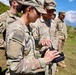 A group of U.S. Army soldiers in MultiCam (OCP) camouflage uniforms are gathered outdoors in a mountainous, green terrain under a partly cloudy sky. A female soldier in the foreground is operating what appears to be a handheld radio or drone controller. The setting suggests a field training exercise or operational activity in a high-altitude environment.