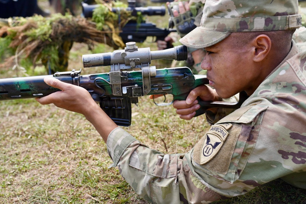 A U.S. Army soldier wearing an 11th Airborne Division patch is in a prone-like position aiming a Soviet/Russian-style rifle with a PSO-1 optical scope, likely during a foreign weapons familiarization training exercise. The weapon appears to be an SVD Dragunov or similar Soviet-era sniper rifle, painted in a camouflage pattern. Additional personnel and weapons are visible in the background, suggesting a group training or demonstration event.
