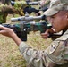 A U.S. Army soldier wearing an 11th Airborne Division patch is in a prone-like position aiming a Soviet/Russian-style rifle with a PSO-1 optical scope, likely during a foreign weapons familiarization training exercise. The weapon appears to be an SVD Dragunov or similar Soviet-era sniper rifle, painted in a camouflage pattern. Additional personnel and weapons are visible in the background, suggesting a group training or demonstration event.