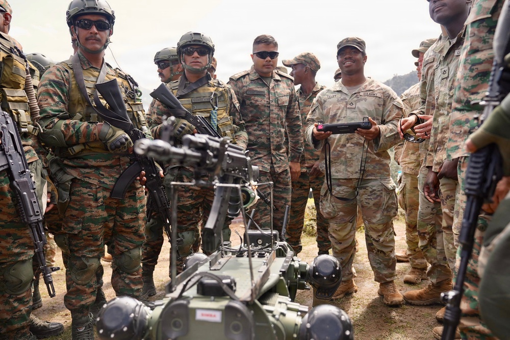 A joint military exercise appears to be taking place between U.S. Army soldiers and Indian Army soldiers. A U.S. Army soldier in MultiCam/OCP uniform is demonstrating a ground robot using a handheld controller to a group of Indian Army soldiers in DPCU (Disruptive Pattern Camouflage Uniform). The setting appears to be an outdoor training area with hilly terrain visible in the background.