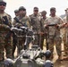A joint military exercise appears to be taking place between U.S. Army soldiers and Indian Army soldiers. A U.S. Army soldier in MultiCam/OCP uniform is demonstrating a ground robot using a handheld controller to a group of Indian Army soldiers in DPCU (Disruptive Pattern Camouflage Uniform). The setting appears to be an outdoor training area with hilly terrain visible in the background.