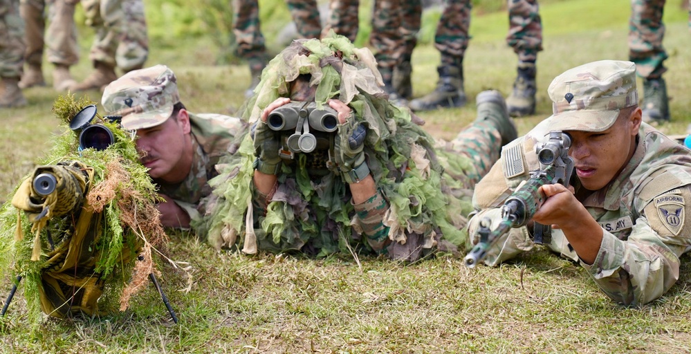 Three military personnel are in a prone position on grass, appearing to conduct a sniper or reconnaissance training exercise. The soldier on the right wears U.S. Army Airborne insignia and aims a rifle, the center figure wears a ghillie suit and uses binoculars, and the left soldier operates a scoped weapon on a bipod. Additional uniformed personnel are visible standing in the background, suggesting an observed training demonstration.