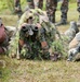 Three military personnel are in a prone position on grass, appearing to conduct a sniper or reconnaissance training exercise. The soldier on the right wears U.S. Army Airborne insignia and aims a rifle, the center figure wears a ghillie suit and uses binoculars, and the left soldier operates a scoped weapon on a bipod. Additional uniformed personnel are visible standing in the background, suggesting an observed training demonstration.